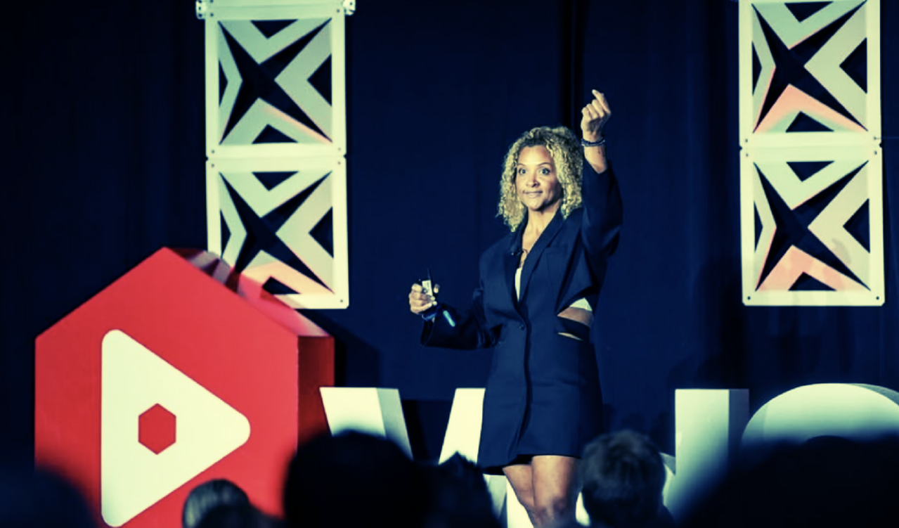 A person giving a presentation on stage, standing near a large red and white logo with an audience visible in the foreground.